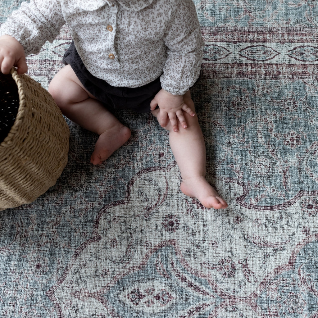 A baby sits on a memory foam playmat rug in deep red and blue-grey tones.