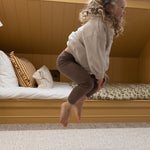 A happy little girl jumps joyfully in her bedroom on a soft playmat with a beautiful Morris & Co. Acorn design