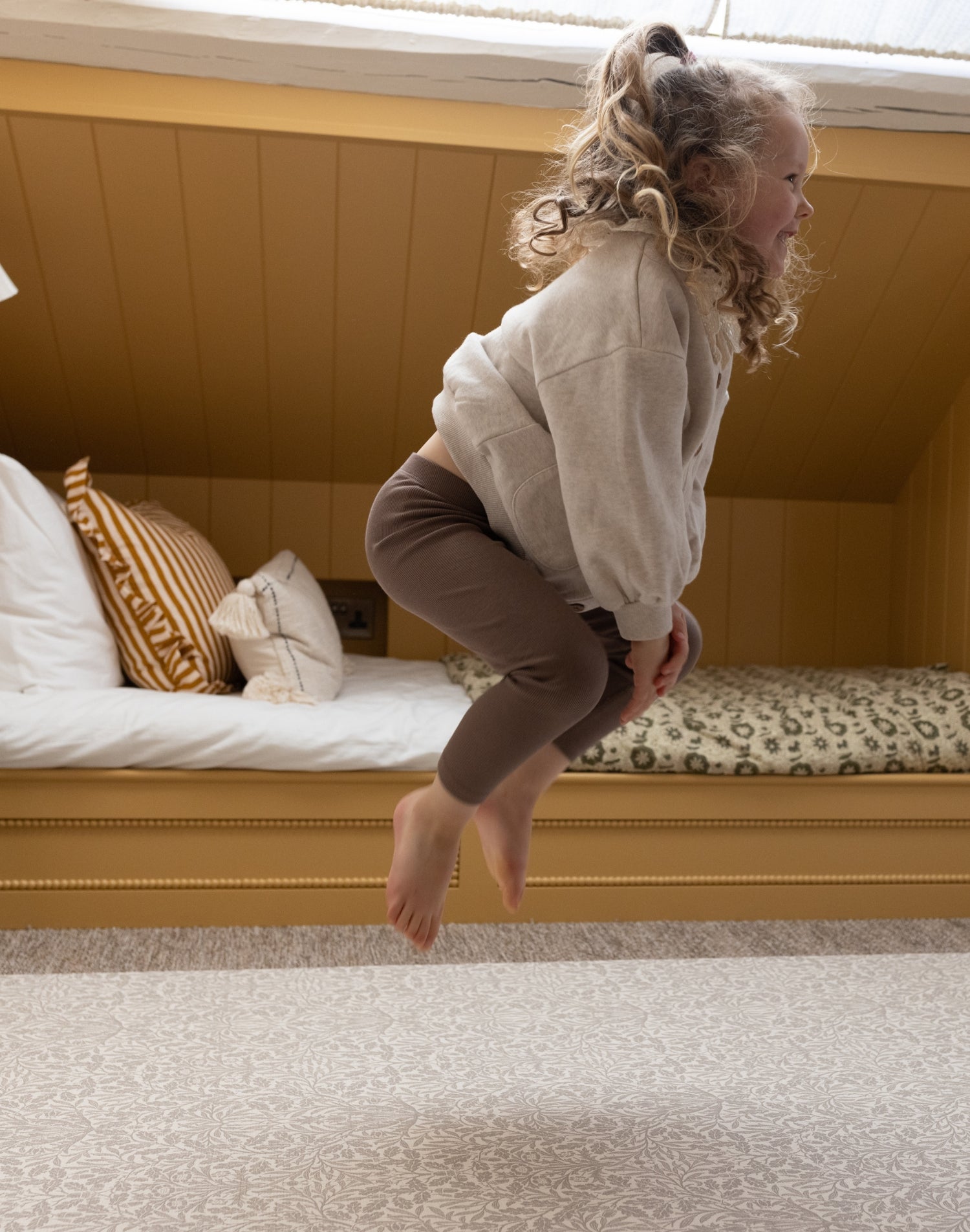 A happy little girl jumps joyfully in her bedroom on a soft playmat with a beautiful Morris & Co. Acorn design