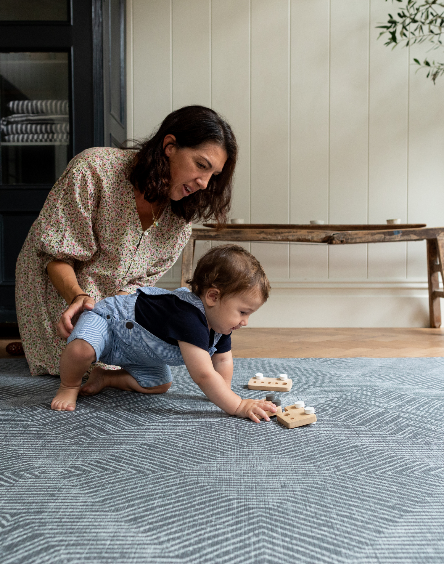xMother places gentle arm on little boy playing with wooden car toys on the Astronomer supportive grey baby play mat