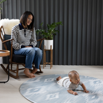 Mother sits in chair watching baby on Large round play mat by Totter and Tumble acts like a wipeable rug in the family home easy to clean spit up and spillages and looks stylish