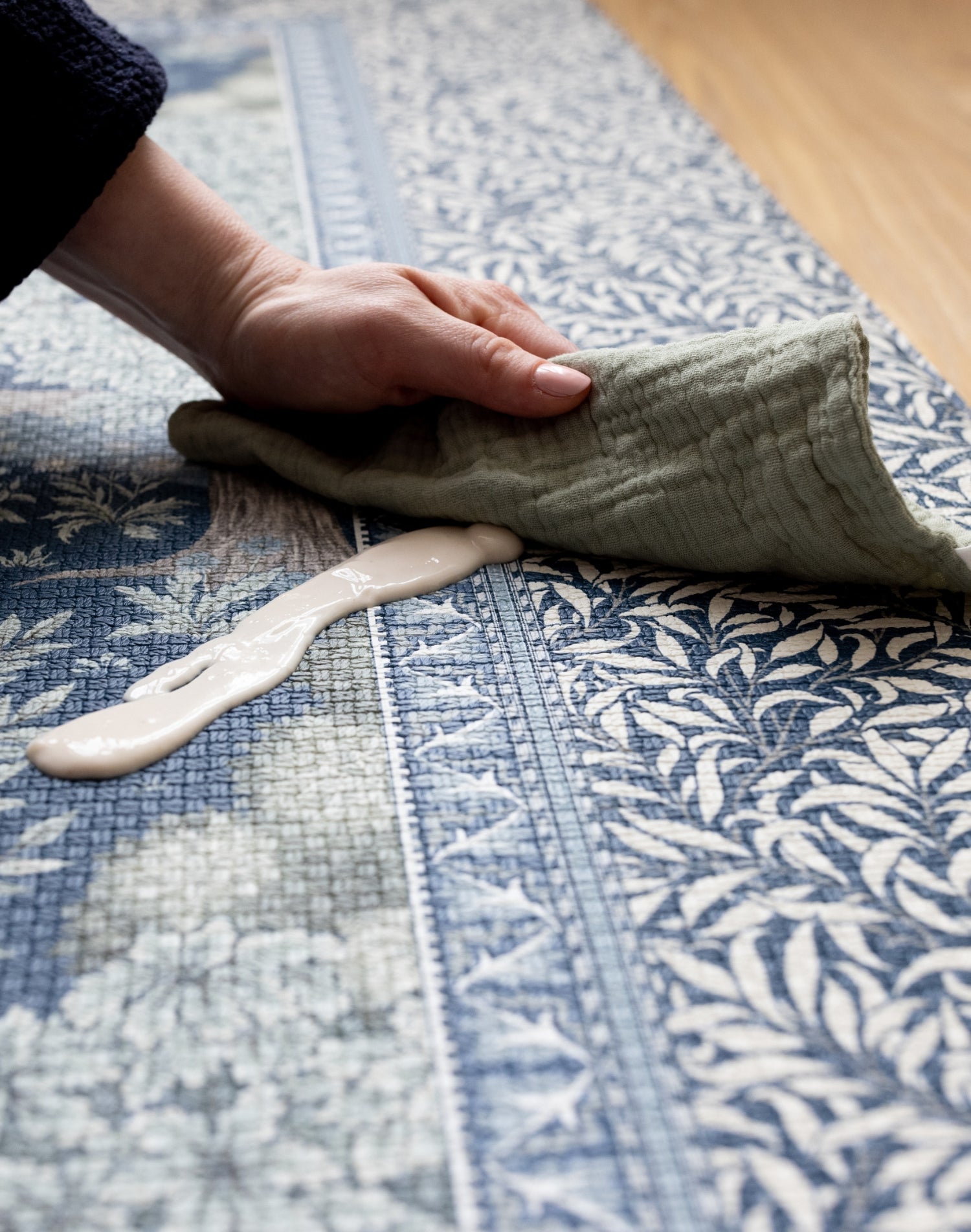 Detailed view of the durable Totter + Tumble nursery Brook play mat with nature-inspired patterns and high-end Morris & Co. craftsmanship. A hand and muslin cloth wipe up a spill on the playmat with ease.