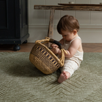 Little one sits supported on an olive green play mat playing with a rustic basket