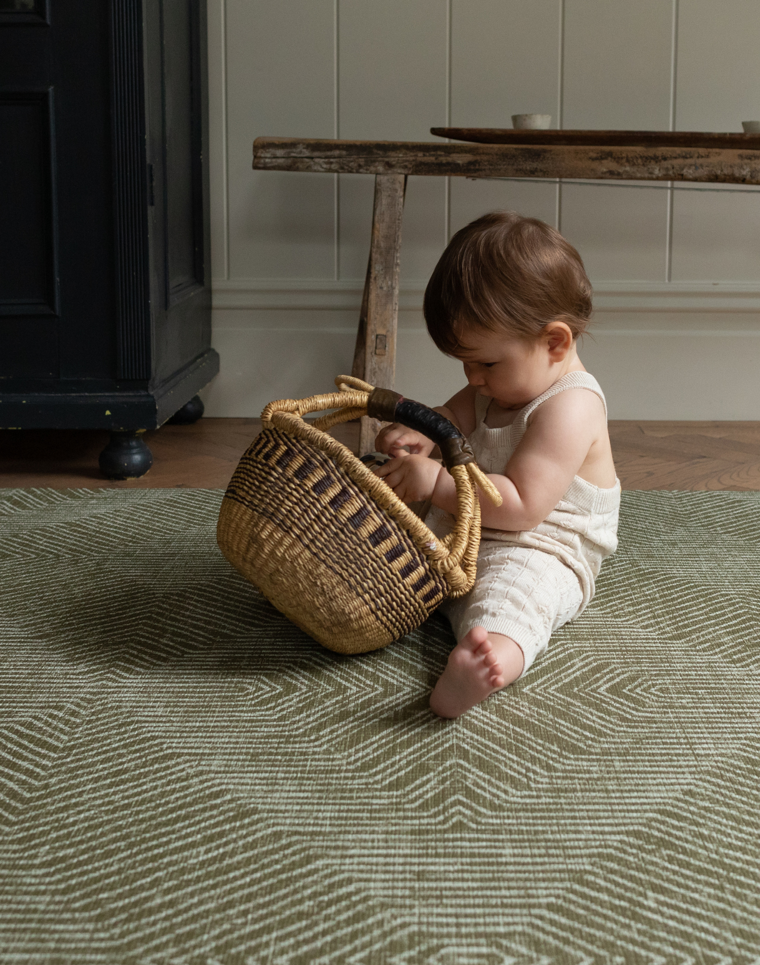 Little one sits supported on an olive green play mat playing with a rustic basket