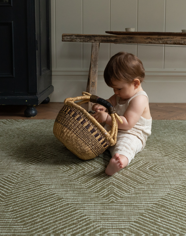 Little one sits supported on an olive green play mat playing with a rustic basket