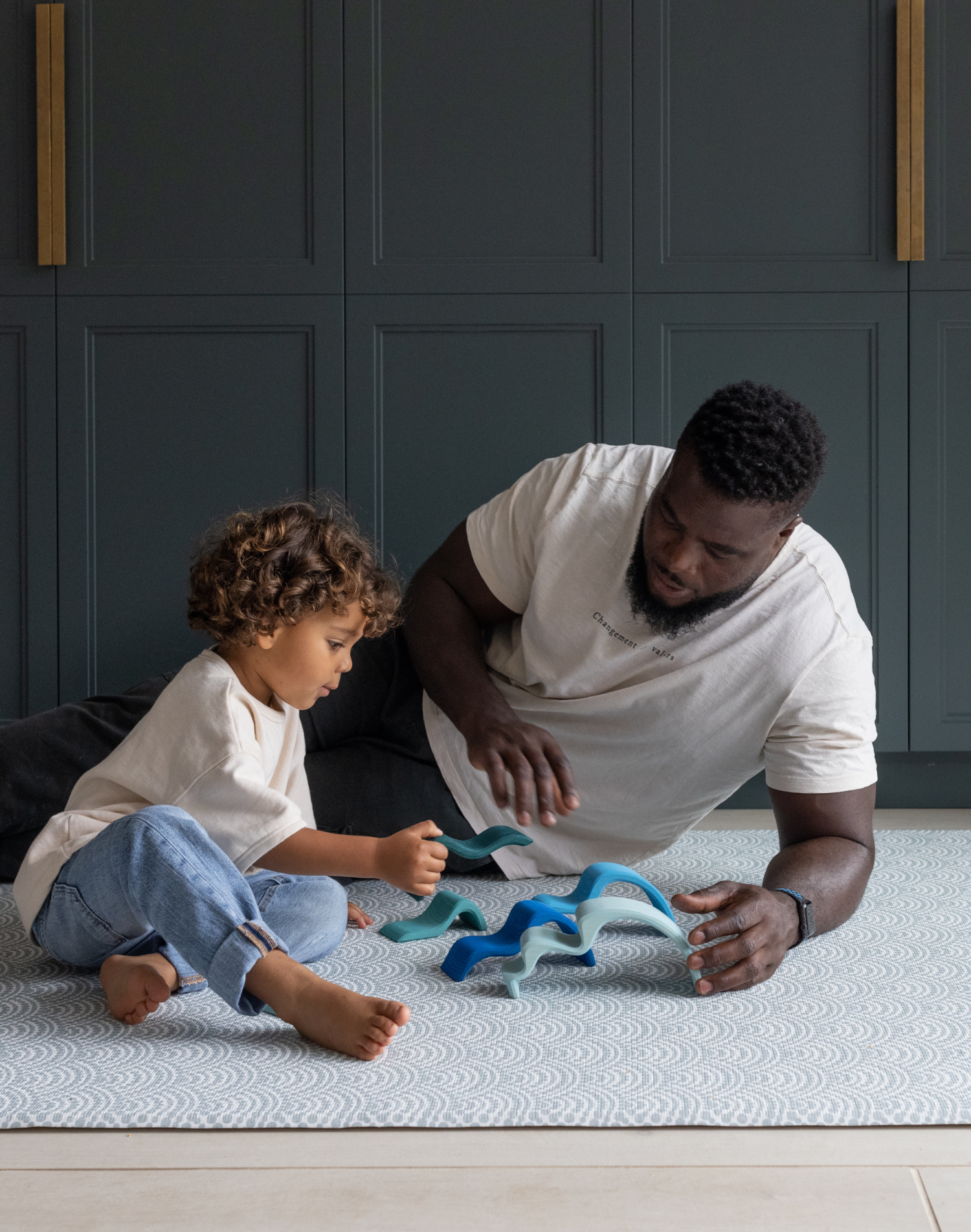 Father and son sitting comfortably on thick play mat designed with memory foam for protection on hard floors playing with wooden Grimms toys