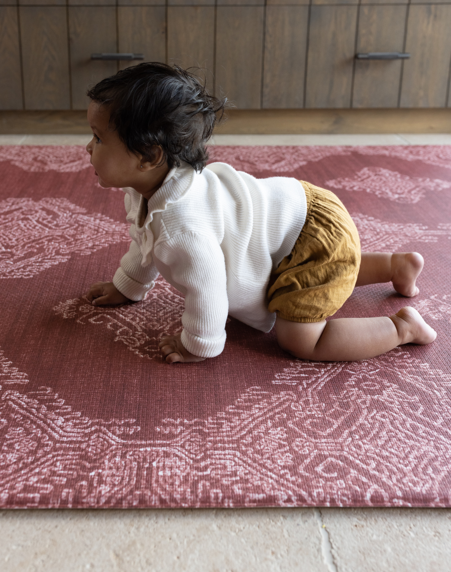 Baby crawling on deep red play mat with intricate batik inspired detailing memory foam provides support for knees and crawling babies