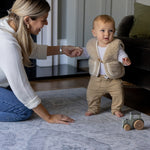 A mother holds the hand of a baby who is learning to walk. The baby is walking on a big soft foam play mat that has a beautiful grey design