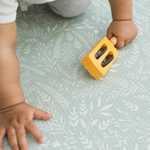 Baby holds a wooden instrument and crawls across a botanical play mat that supports movement with comfy memory foam