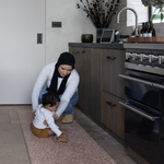 Baby sits on the orange kitchen mat The Rusa providing protection for little ones in the kitchen while also allowing parents to keep comfortable during household chores