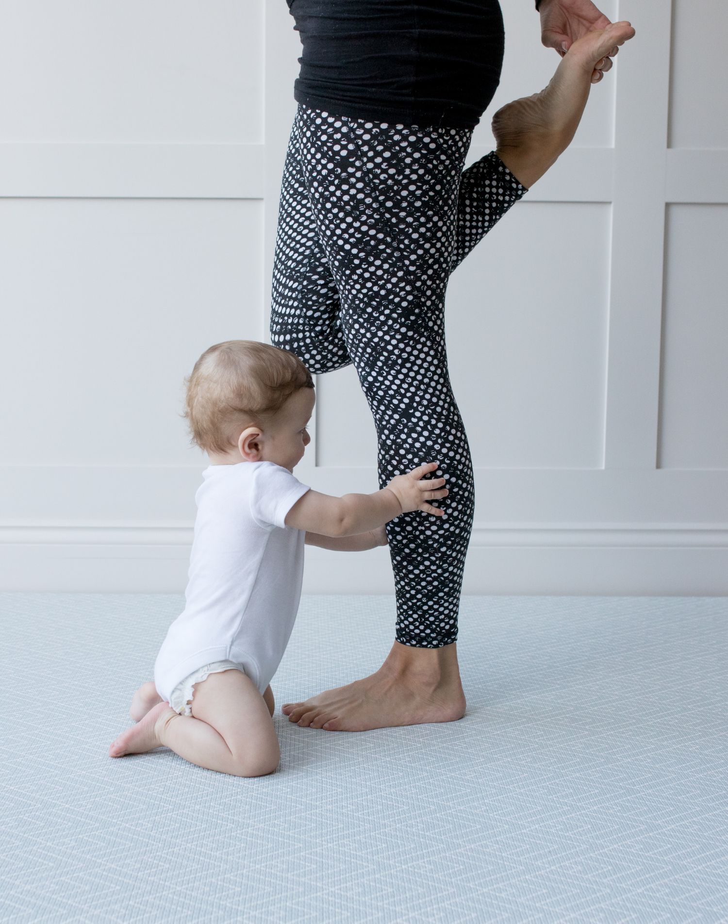 Little baby holds the legs of lady working out on the large grey exercise mat by totter and tumble