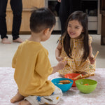 Two children sitting on the floor in a kitchen, eating snacks from colorful bowls on a muted easy to clean playmat from Totter and Tumble