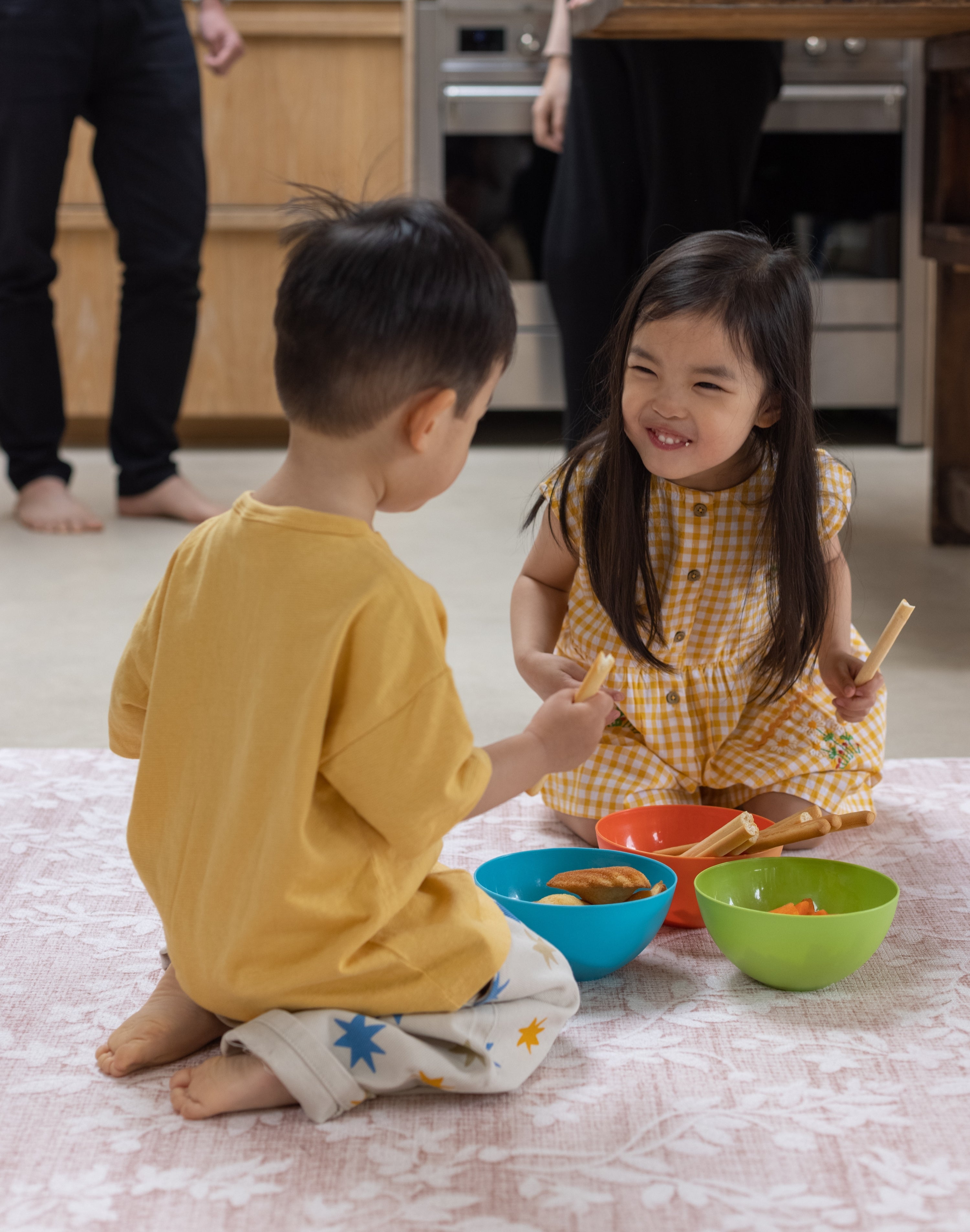 Two children sitting on the floor in a kitchen, eating snacks from colorful bowls on a muted easy to clean playmat from Totter and Tumble