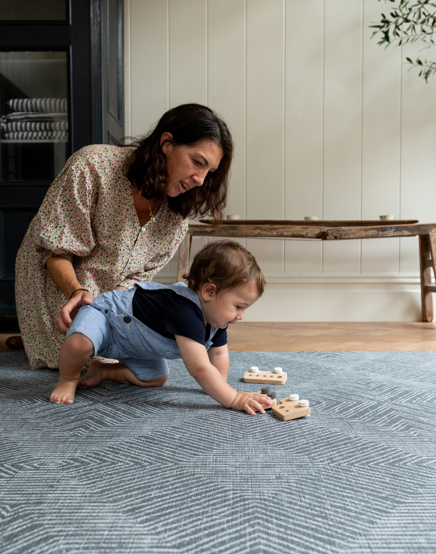 xMother places gentle arm on little boy playing with wooden car toys on the Astronomer supportive grey baby play mat 