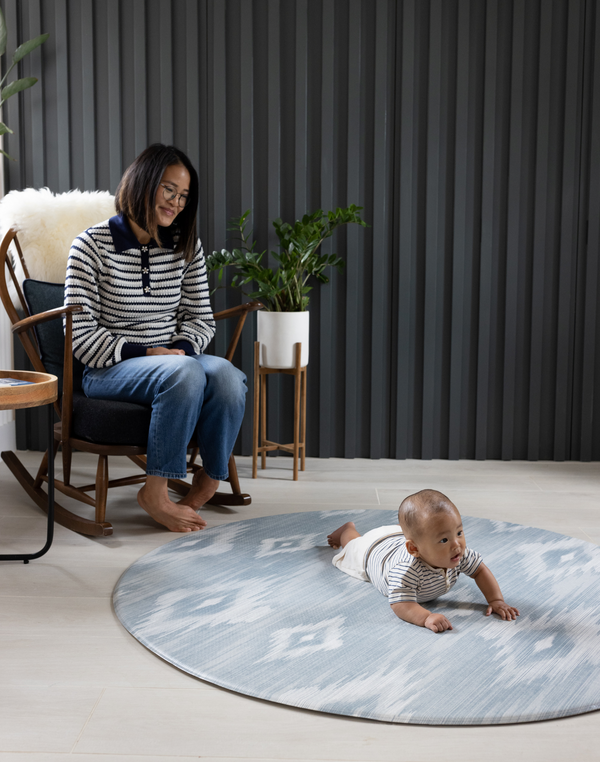 Mother sits in chair watching baby on Large round play mat by Totter and Tumble acts like a wipeable rug in the family home easy to clean spit up and spillages and looks stylish