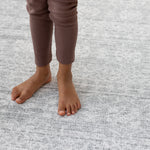 A child's feet stand on a warm large playroom rug. The playmat has a beautiful grey pattern