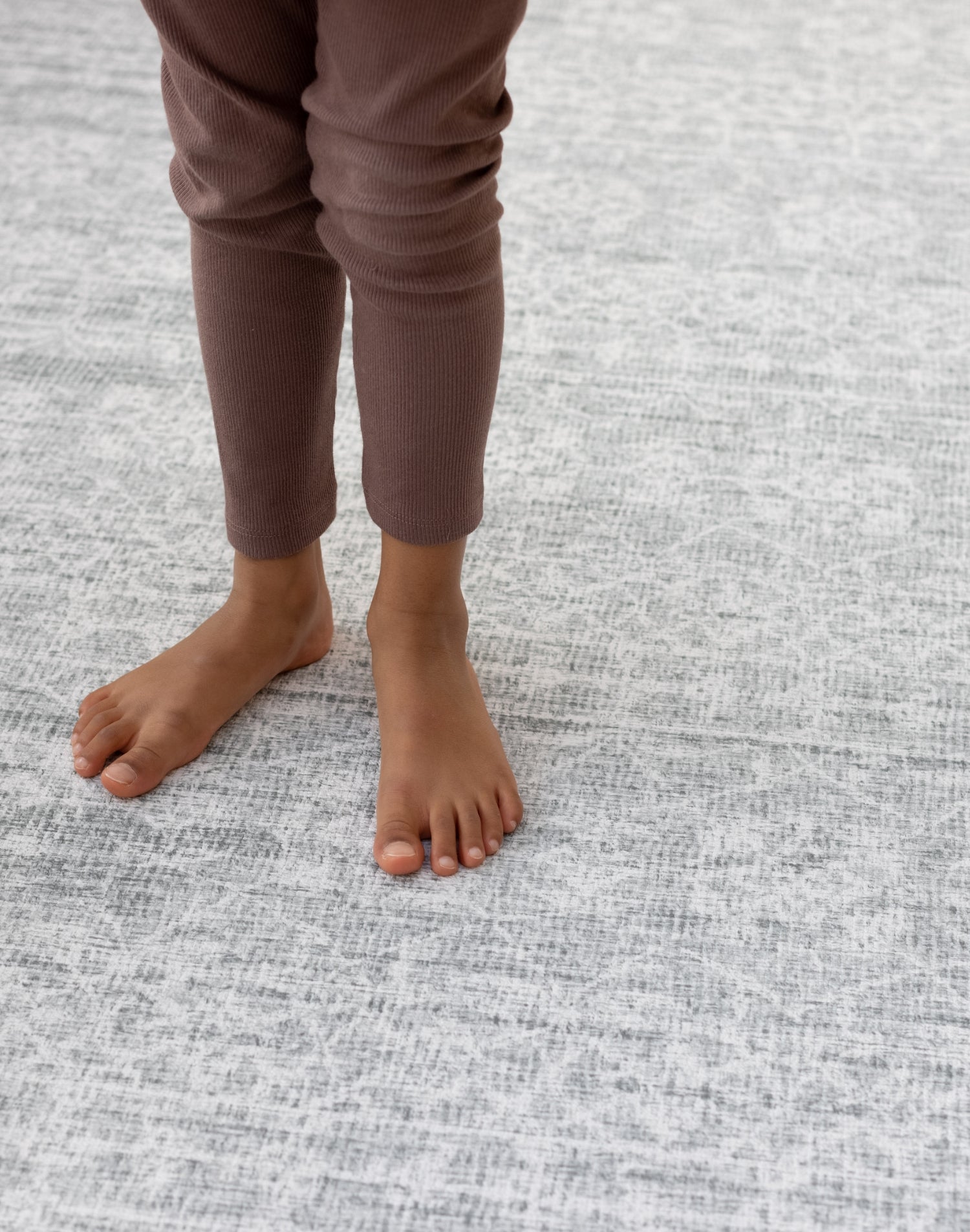 A child's feet stand on a warm large playroom rug. The playmat has a beautiful grey pattern