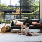 A family room scene. A toddler plays with stacking blocks while a baby lies down looking comfortable. They are on a large grey patterned playmat for infants