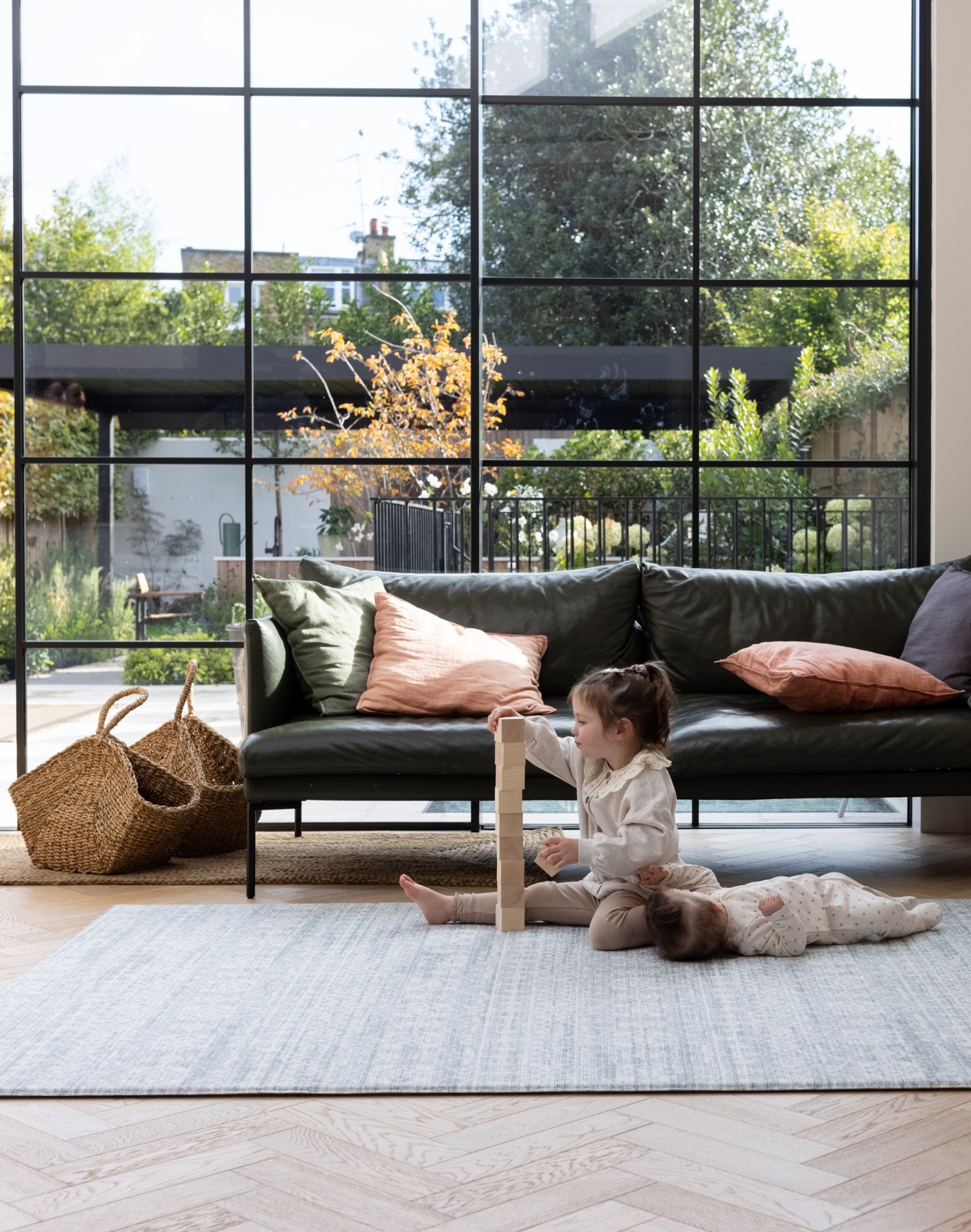 A family room scene. A toddler plays with stacking blocks while a baby lies down looking comfortable. They are on a large grey patterned playmat for infants