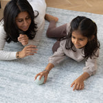 A mother and daughter are dressed in neutral tones. They lie down and play together with a ball on a big soft play mat