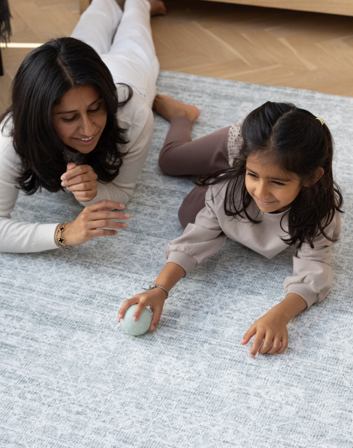 A mother and daughter are dressed in neutral tones. They lie down and play together with a ball on a big soft play mat