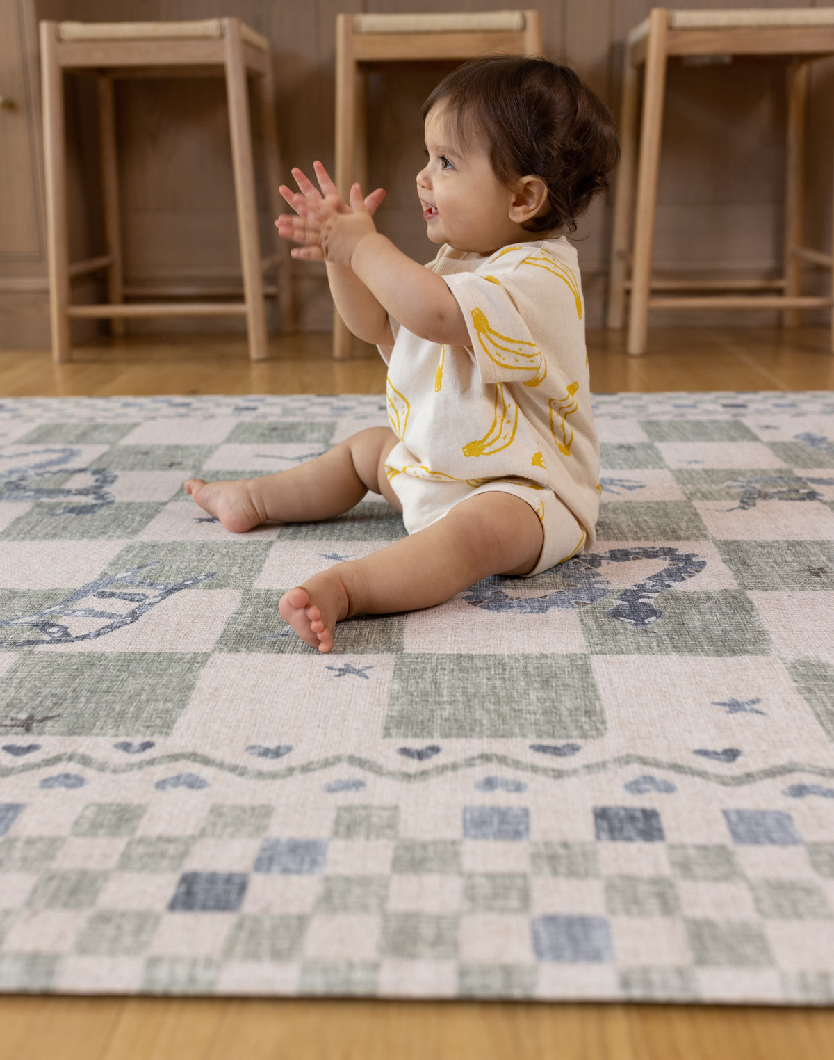 Baby sitting on a check snakes and ladders fun design one piece playmat looks like a rug in a room with wooden furniture.
