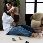 Lady holds baby and sits comfortably on soft baby playmat designed for the whole family 