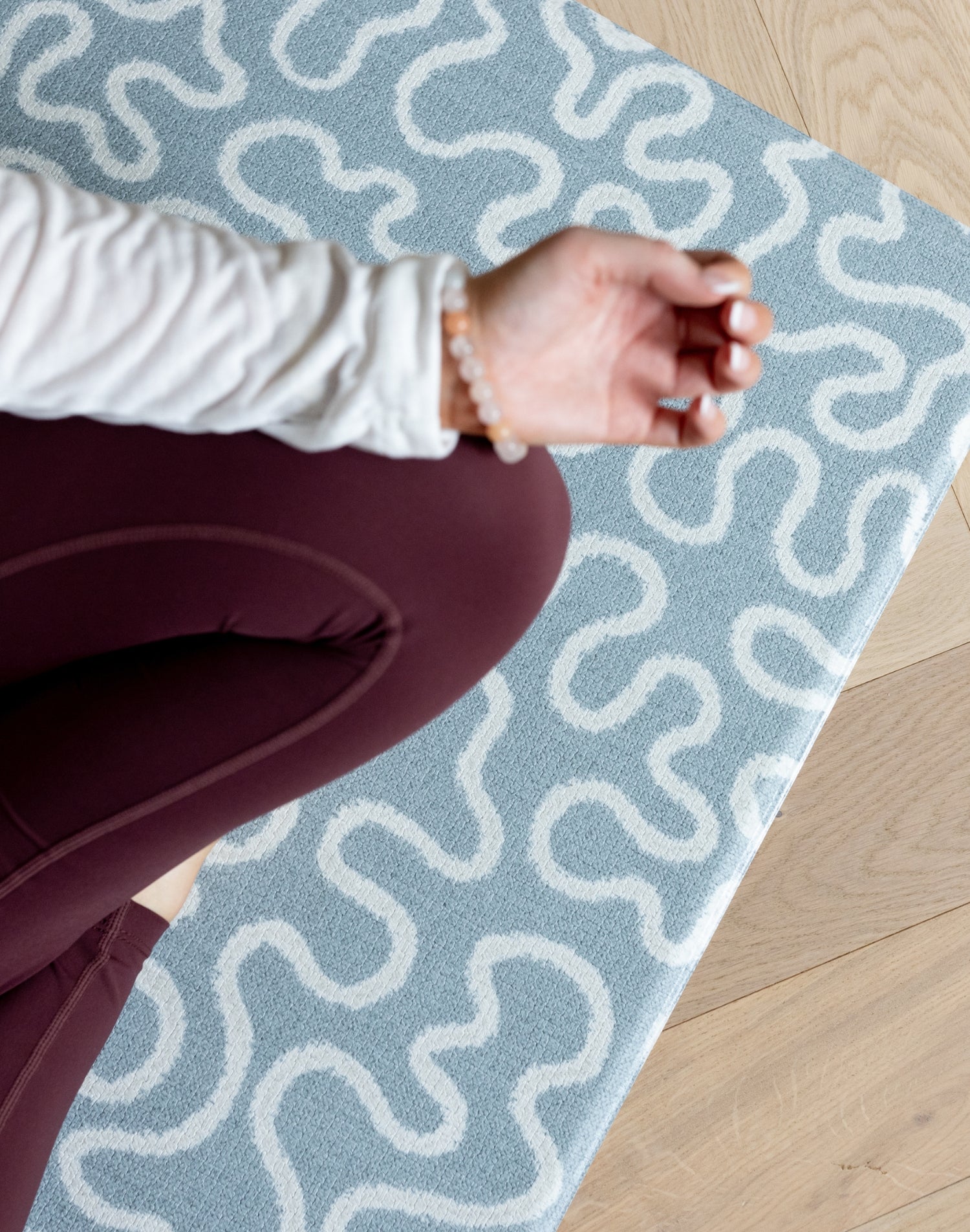 Person sitting on a blue and white patterned yoga mat on a wooden floor.