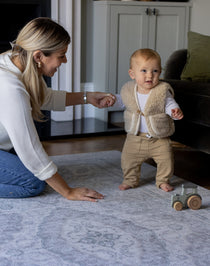 A mother holds the hand of a baby who is learning to walk. The baby is walking on a big soft foam play mat that has a beautiful grey design