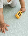 Baby holds a wooden instrument and crawls across a botanical play mat that supports movement with comfy memory foam