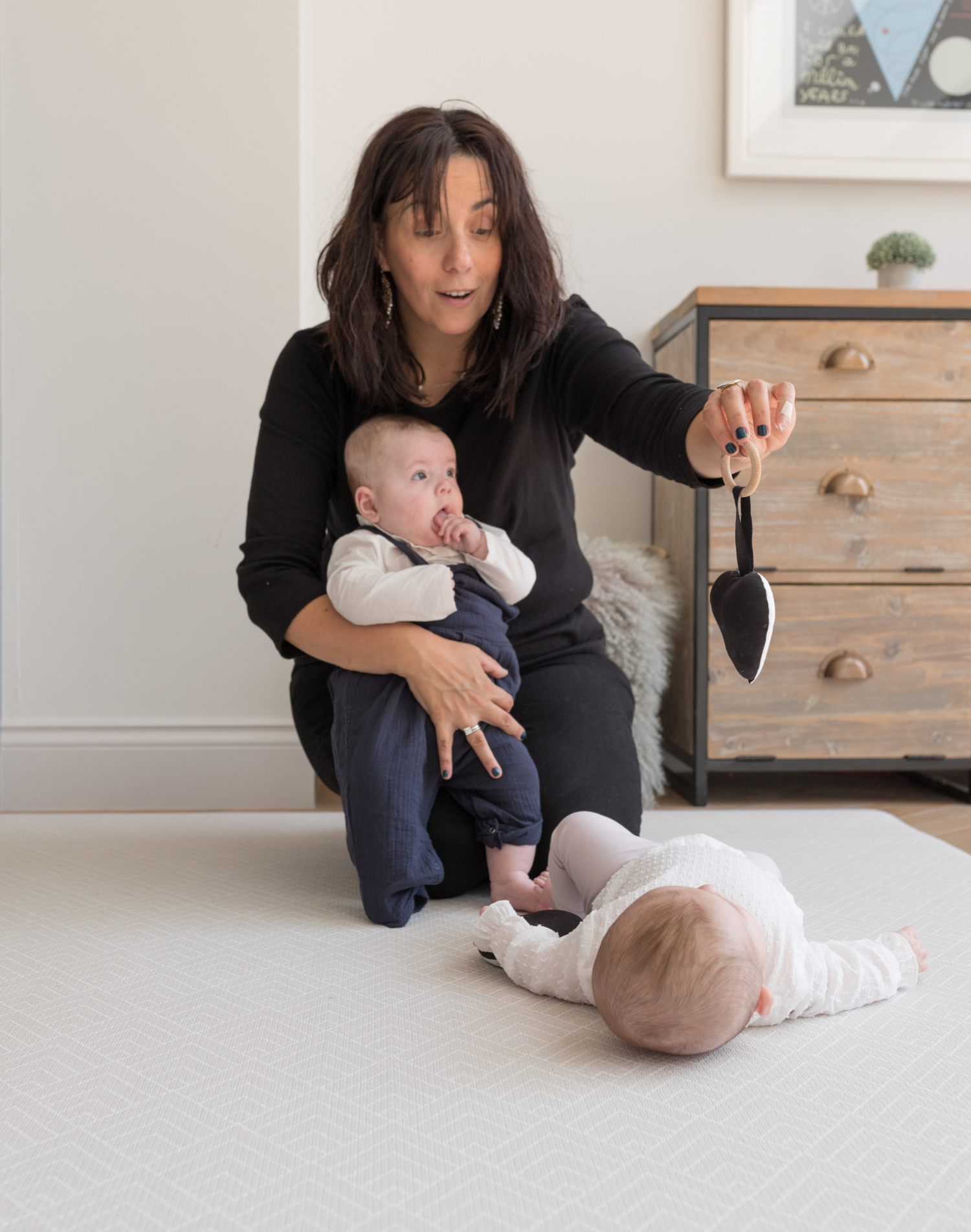 Lady holding one baby and playing with a second baby laying on a thick foam play mat that supports children and adults on the floor