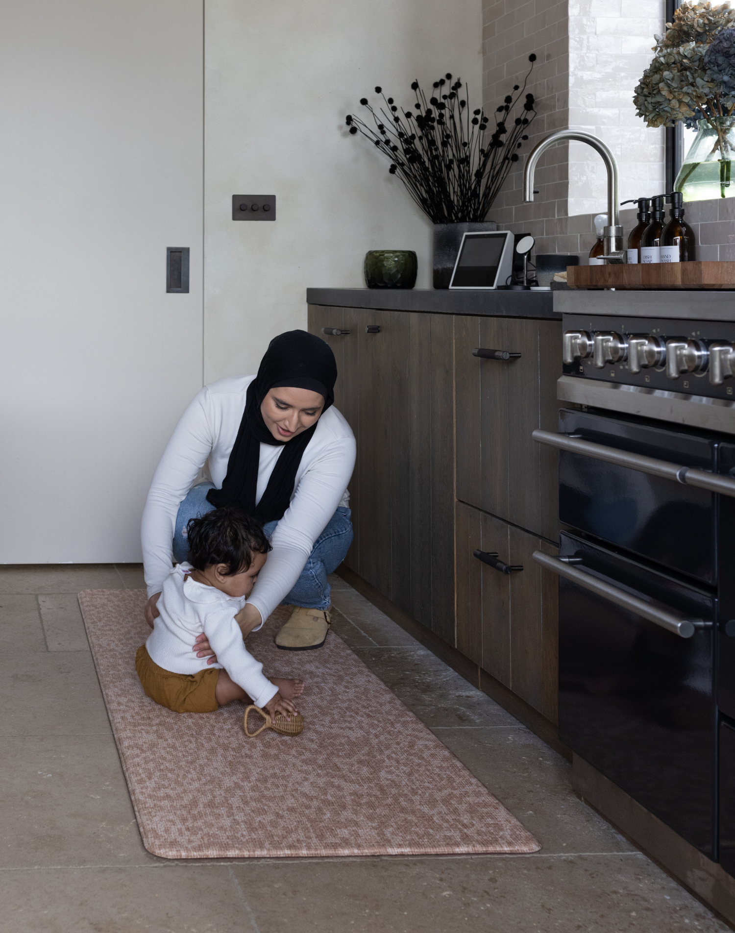 Baby sits on the orange kitchen mat The Rusa providing protection for little ones in the kitchen while also allowing parents to keep comfortable during household chores