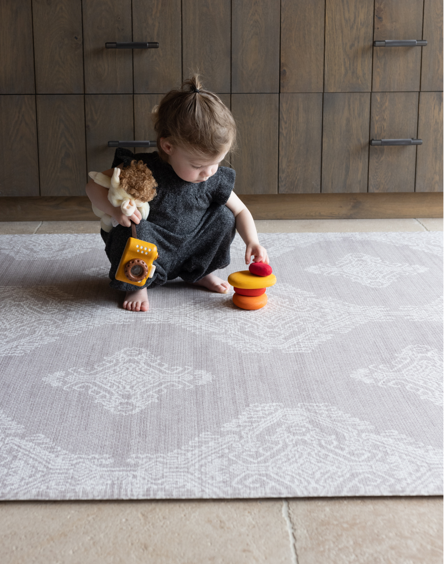 Toddler stacking smooth wooden pebbles and playing with other toys on the soft Grey play mat the Sepi with a delicate batik design that looks like a rug thick memory foam supports as she enjoys floortime