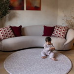 Baby girl plays with toy sitting on animal print play mat in light grey tone small round play mat size is ideal for placing in front of sofa for safe play in the family home