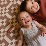 Two children lying down  and smiling on a brown and red patterned playmat from Totter + Tumble