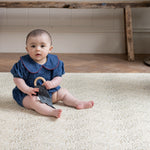 Baby sitting on a patterned playmate holding a toy in a room with wooden floor and white wall.