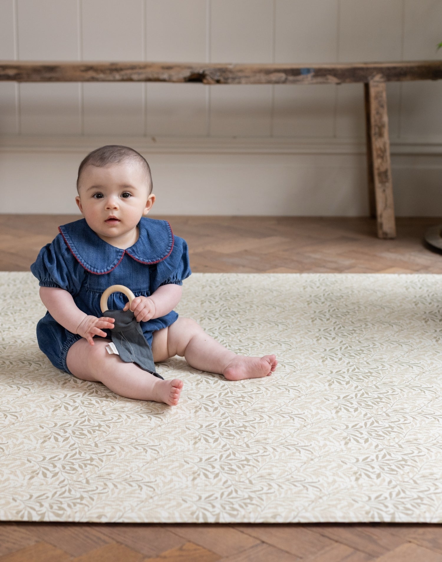 Baby sitting on a patterned playmate holding a toy in a room with wooden floor and white wall.