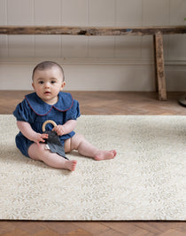 Baby sitting on a patterned playmate holding a toy in a room with wooden floor and white wall.