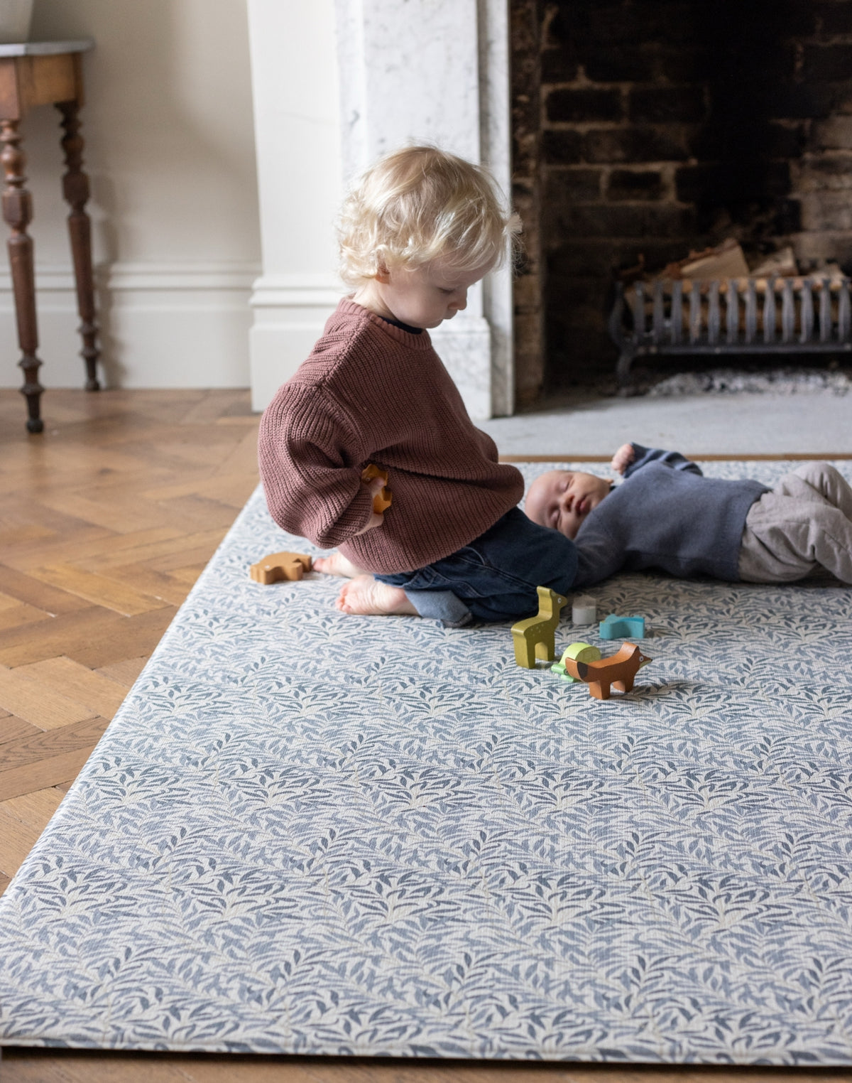 Toddler and baby playing with wooden toys on Totter and Tumble Morris & Co. Willow Boughs Woad Playmat reversible memory foam playmat against fireplace