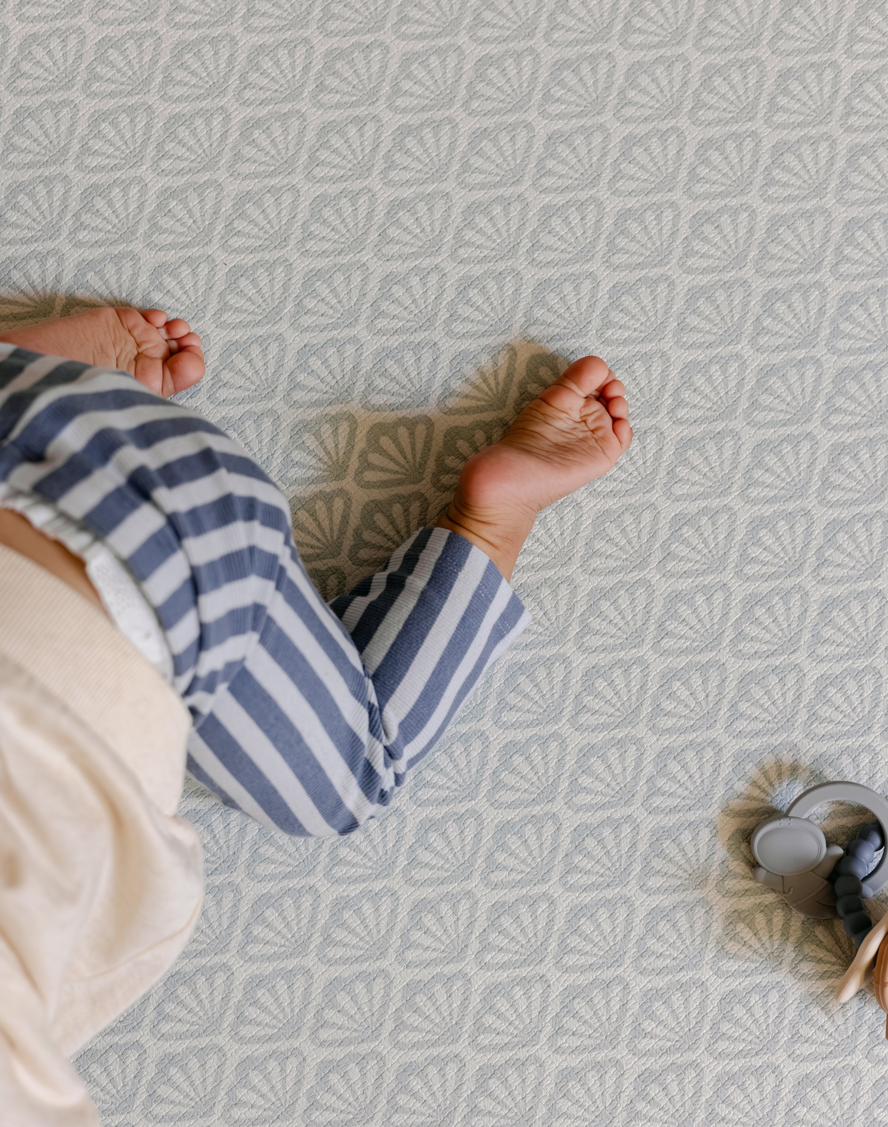 A baby's bare feet and legs wearing blue and white striped pants, crawling on a shell patterned totter and tumble playmat