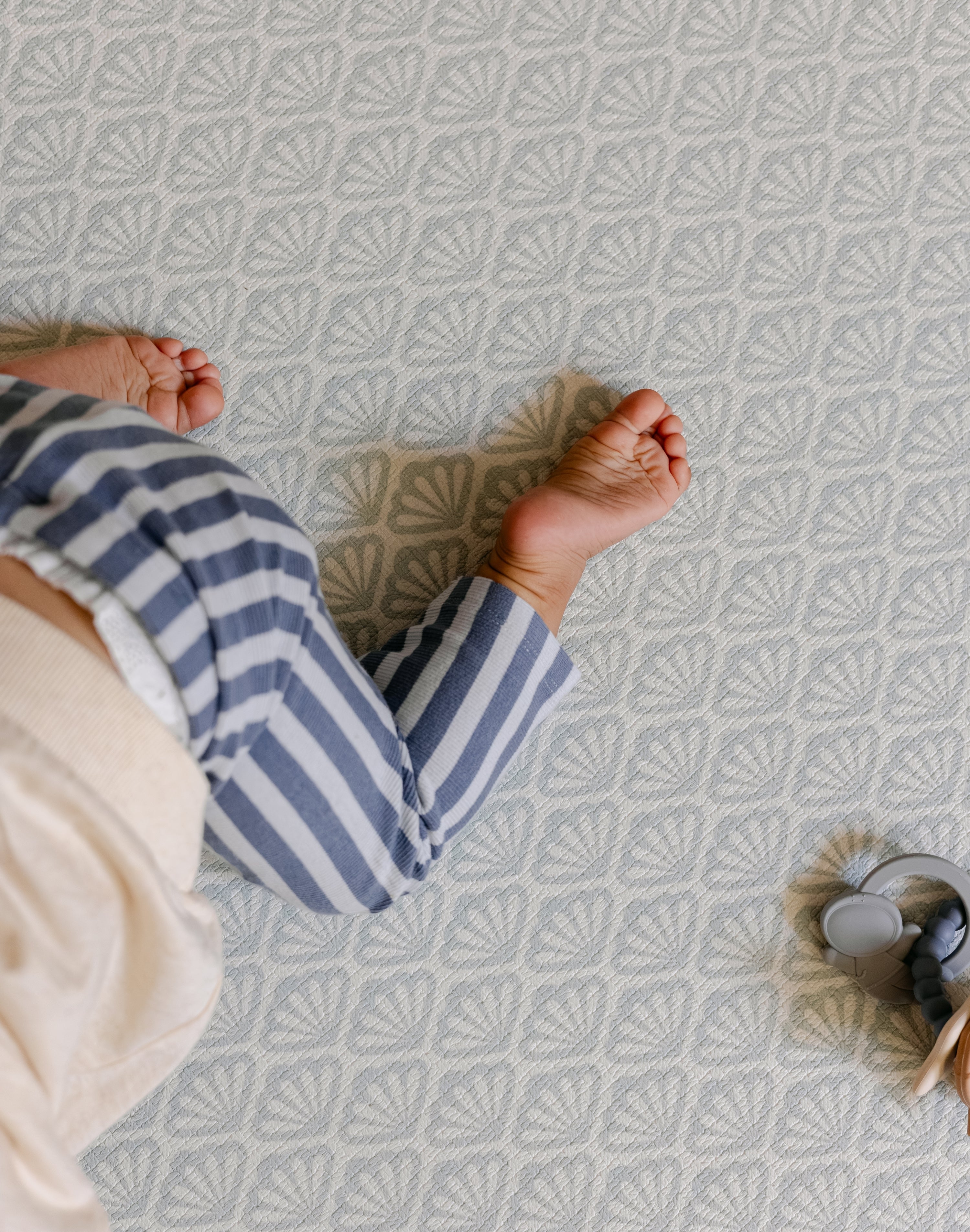 A baby's bare feet and legs wearing blue and white striped pants, crawling on a shell patterned totter and tumble playmat