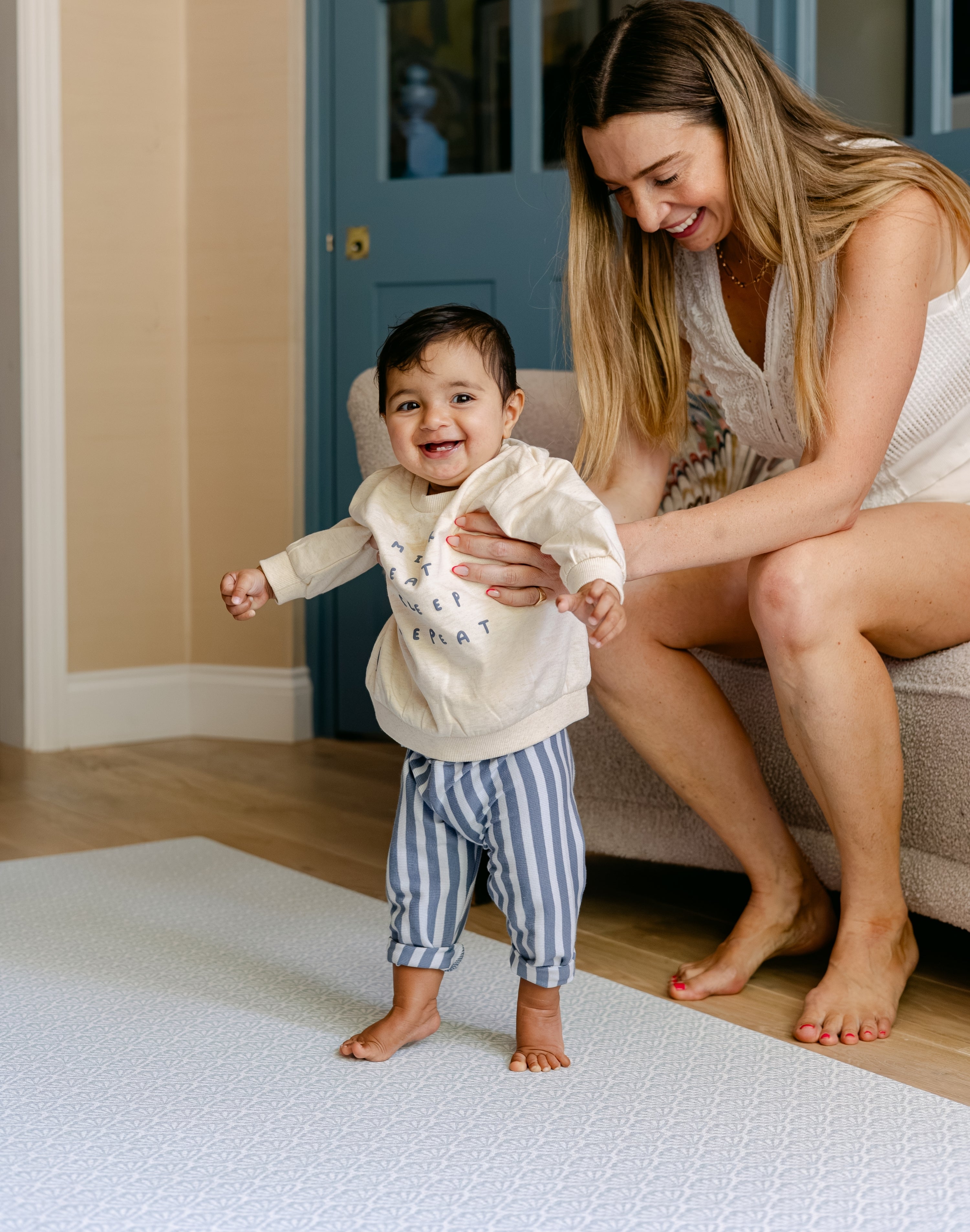A baby standing up with help from it's mother safe and comfortably on a thick totter and tumble memory foam playmat