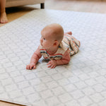 A baby wearing a striped romper lies on their stomach on a light geometric patterned totter and tumble The Gatherer baby play mat in a living room setting.