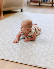 A baby wearing a striped romper lies on their stomach on a light geometric patterned totter and tumble The Gatherer baby play mat in a living room setting.