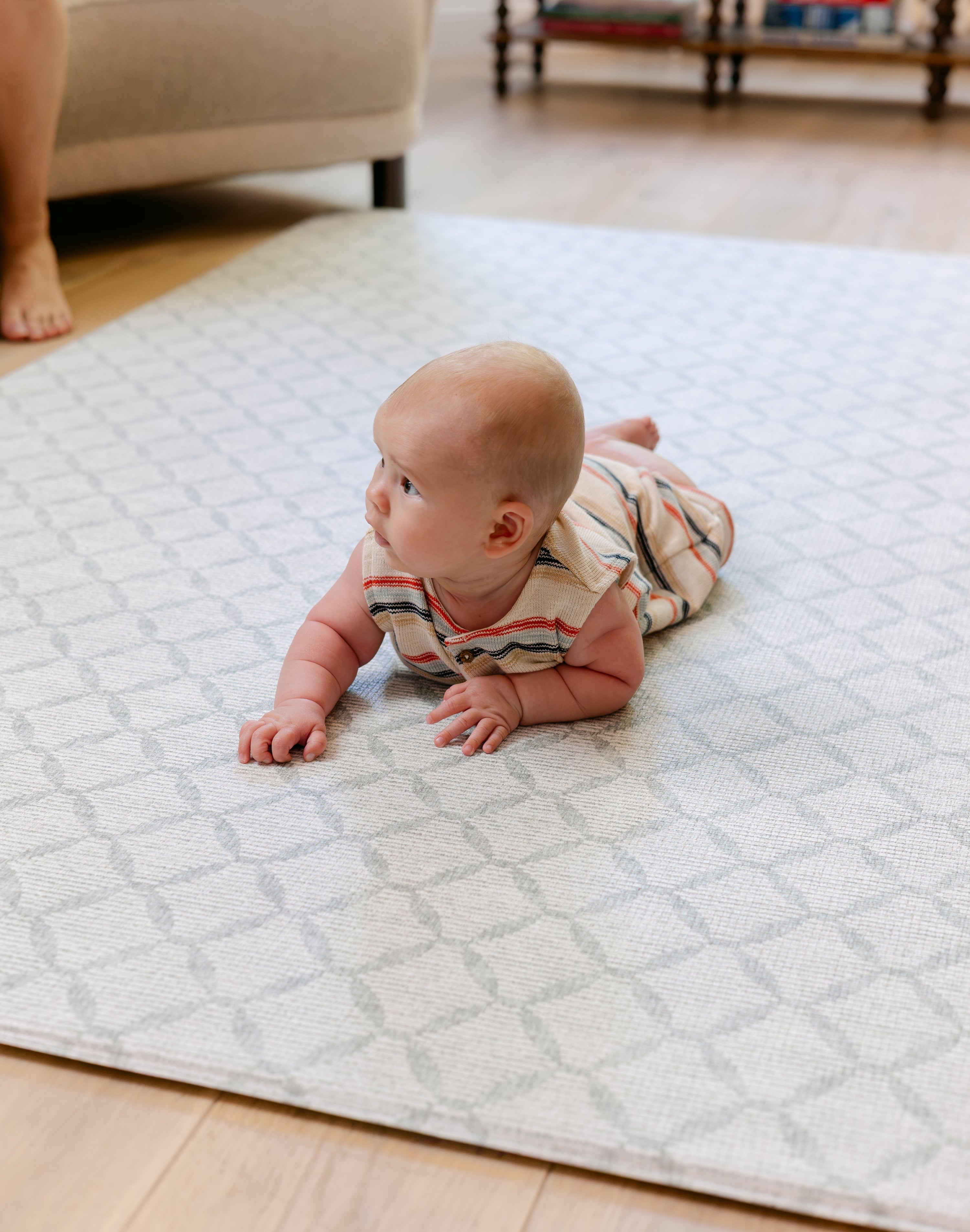 A baby wearing a striped romper lies on their stomach on a light geometric patterned totter and tumble The Gatherer baby play mat in a living room setting.