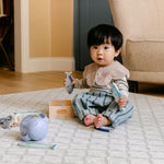 A toddler sits barefoot on a patterned totter and tumble 'the gatherer' playmat in a living room, holding two light blue toy fish. 