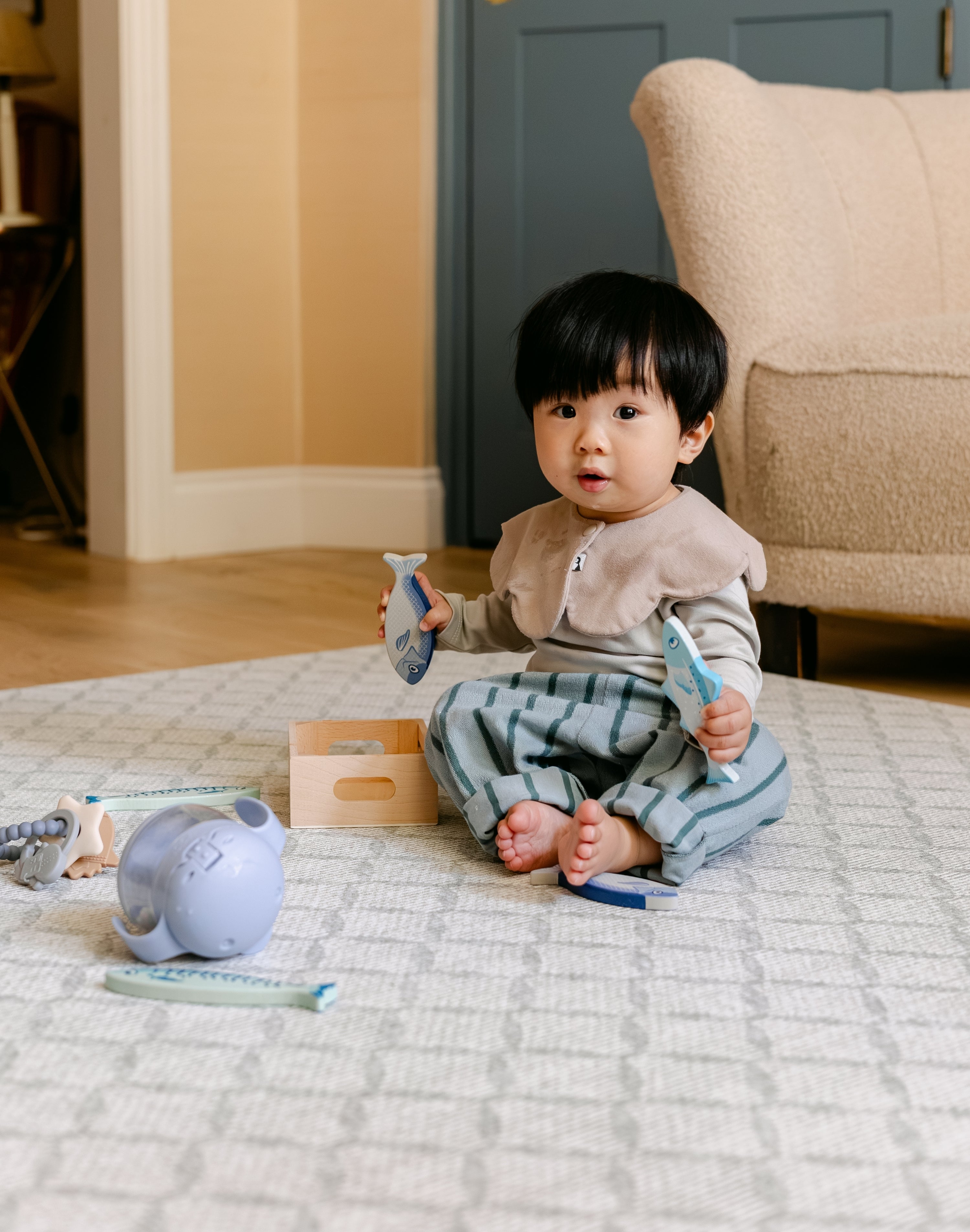 A toddler sits barefoot on a patterned totter and tumble 'the gatherer' playmat in a living room, holding two light blue toy fish. 