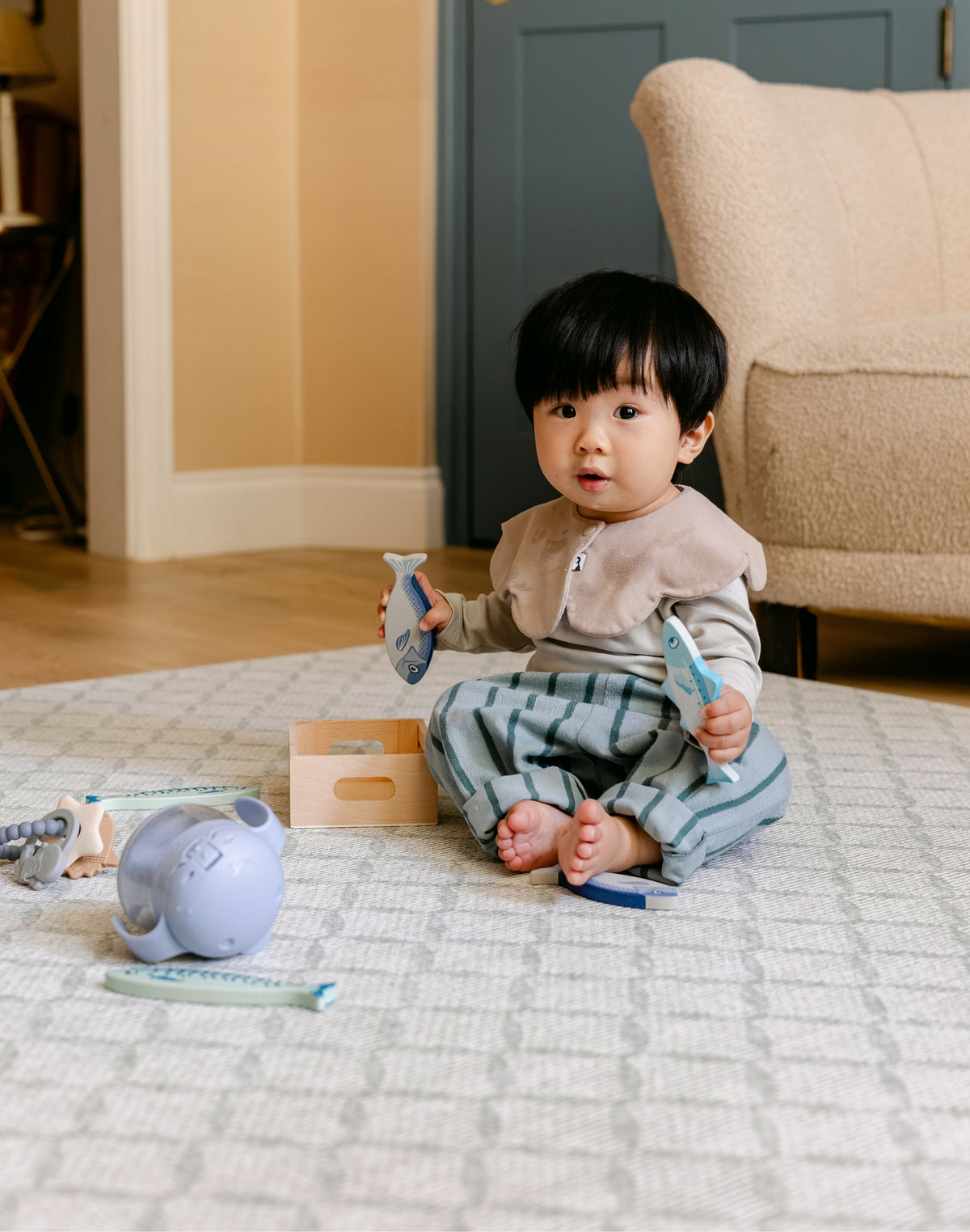 A toddler sits barefoot on a patterned totter and tumble 'the gatherer' playmat in a living room, holding two light blue toy fish. | SKU: 26 BEACH/GATHER XL, 26 BEACH/GATHER STD, 26 BEACH/GATHER COMP