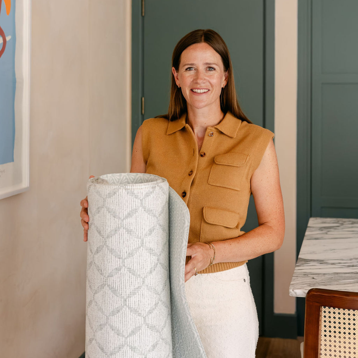 Susie, the founder of totter and tumble stands holding a rolled the gatherer and the beachcomber reversible playmat. 