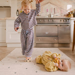 Toddler balancing in the kitchen on a The Wildcard Totter + Tumble reversible wipeable memory foam playmat while the baby lies on it